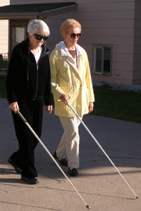 two older white women wearing sunglasses walking together, both using white canes