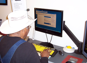Older man using computer with large print keyboard, track ball mouse, and adjustable arm on monitor