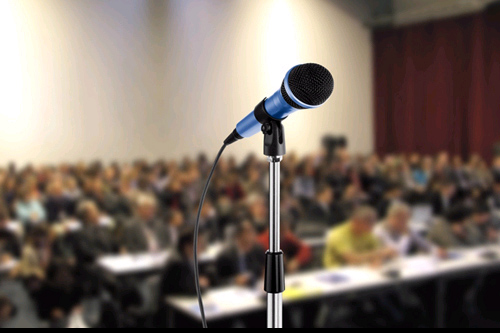 closeup of a microphone stand, ready to be used. In the background, an audience waits to be addressed.