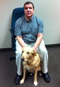 Tim McIsaac sitting in a chair with his guide dog, Jerry, sitting in front