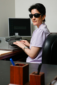woman wearing sunglasses sitting at desk, with computer and braille writer in front of her