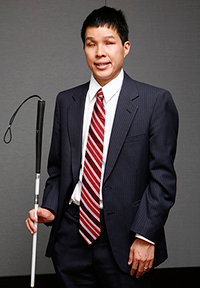 Asian man wearing a gray suit, smiling and holding white cane