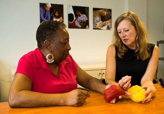 Two women seated while one of them demonstrates the difference between muscle tissue and fat using tactile examples