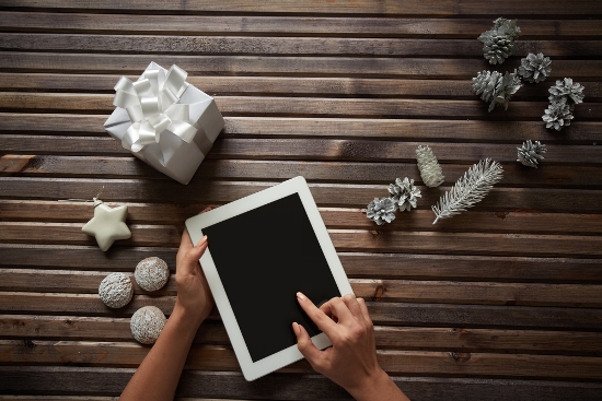 Three spice-cakes, decorative silver cones, giftbox and white toy star surrounding female hands touching display of digital tablet .