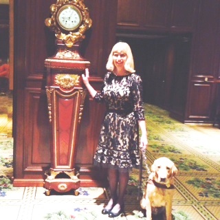 Nancy Shugart and dog guide, Porsche, standing next to a grandfather clock