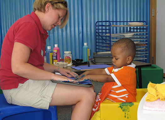 visually impaired little boy enjoying finger painting, with a teacher helping