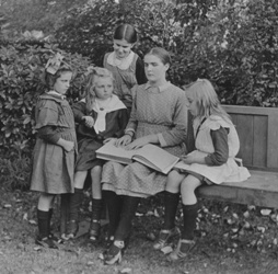 Tilly Aston in her 20s, sitting outside on a bench reading braille and surrounded by small children; Image courtesy of Vision Australia