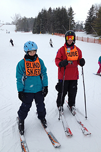 Alyssa on the slopes, wearing her BLIND SKIER vest, with a guide standing next to her