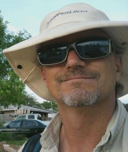 Stephen is sporting a wide brim hat, whit in color with dark sunglasses. Also has a very short cropped mustache and chin beard salt and pepper in color.