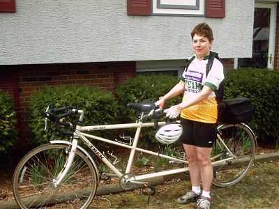 Mary Hiland wearing her biking clothes standing in front of her tandem bike and smiling at the camera