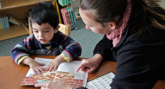 young boy reading a print/braille book as a volunteer looks on