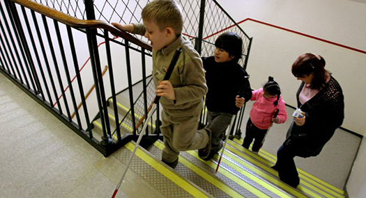 three young children using white canes to go up a school stairway, trailed by an orientation and mobility instructor