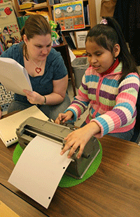 girl using braillewriter in the classroom, with her teacher looking on