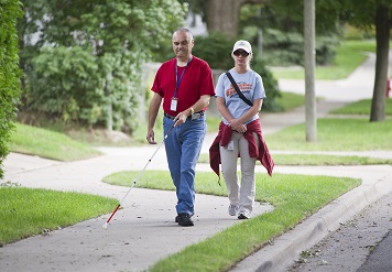 client walking on sidewalk using cane being supervised by instructor