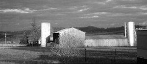 Photo of a farm with a silo at each end of a barn. A fenced field is in the foreground and a mountain range in the background