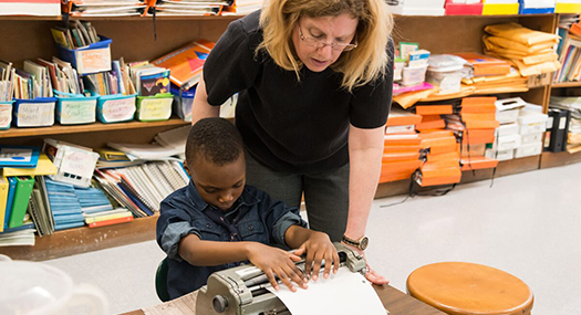 A teacher leans over her student to check his work on the braille embosser