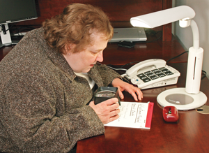 Mujer sentada en  escritorio usando una lupa para leer una libreta. Una lámpara de mesa brillante enciende el  todo el escritorio.