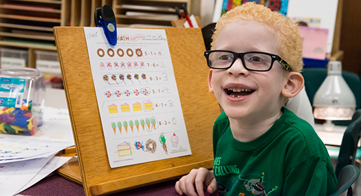 child wearing glasses, smiling with pride next to his large-print math worksheet