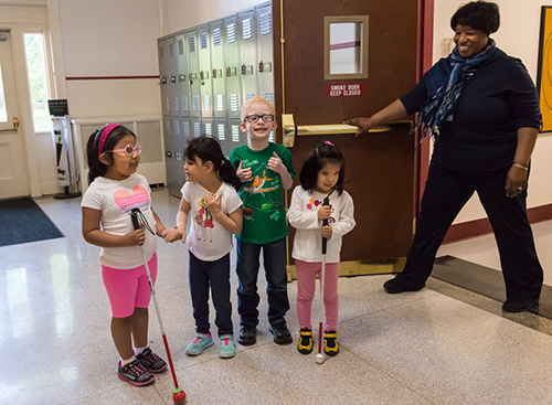 four children, two with canes, two without, smiling as their     orientation and mobility instructor holds the door open for them