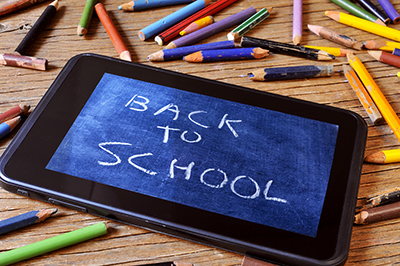 A tablet with a picture of a chalkboard with the sentence back to school written in it, on a rustic wooden desk with worn pencil crayons of different colors 