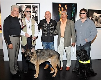 a group of white men gathered in an art gallery - two of them have guide dogs