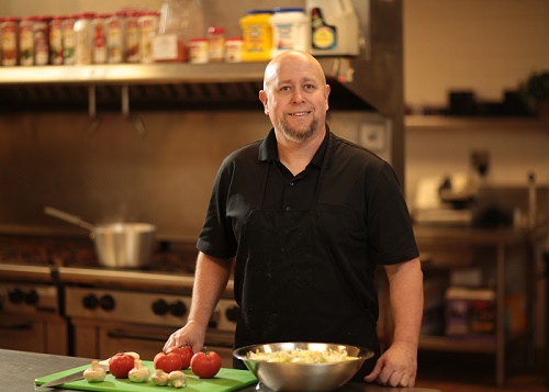 man standing in kitchen with hands on cutting board with vegetables and metal bowl