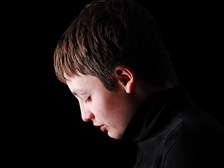 Teenage boy is photographed in profile on the black background. He is upset and his head is hung. 