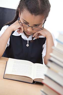 A young girl reading in a school classroom with a pile of books in front of her