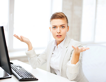 Businesswoman sitting at a computer looking confused with her arms bent up at the elbow