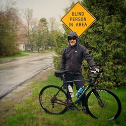 Mike Robertson from Shared Vision Quest standing outside with this bike in front of a road sign stating blind person in area