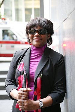 A woman standing outside with her white cane next to a building smiling at the camera