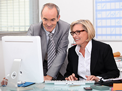 An older man and woman at a desk in business attire working on a computer together