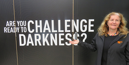 Maribel standing in front of a black wall with the words Are You Ready to Challenge Darkness?