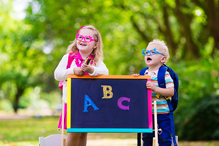 Two children with glasses wearing backpacks in front of a chalkboard with the letters A, B, and C