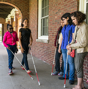 A group of high school students standing outside against a brick wall with two students walking towards the group using white canes