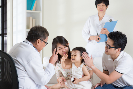 A mother and father holding their small child during an eye examination at the doctor's office; the doctor is looking at the child and holding up three fingers for the baby to see
