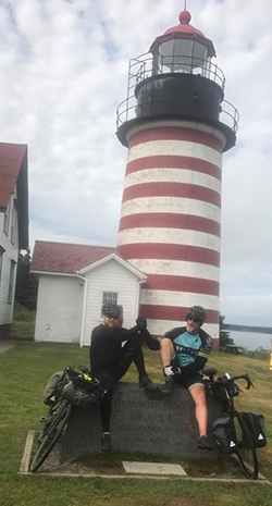 Mike Robertson and his riding partner standing in front of a lighthouse on the coast with their bikes on the ground in front of them