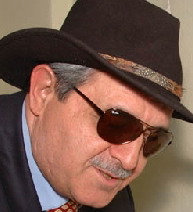 A photo of the painter Esref Armagan. He is wearing sunglasses, a navy blue suit, a red necktie, and a wide-brimmed brown dress hat. Looking good! Credit: Esref Armagan