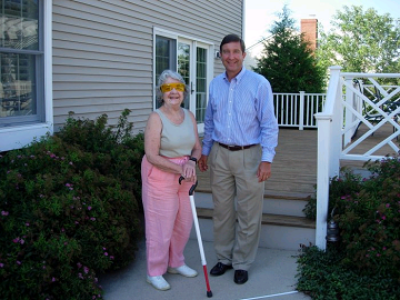 Jane, holding white cane and standing in front of home with son Ed
