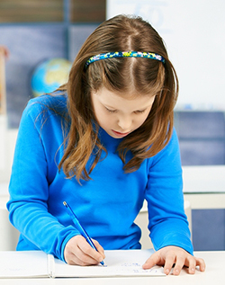 An elementary school age girl in a blue shirt writing at her desk